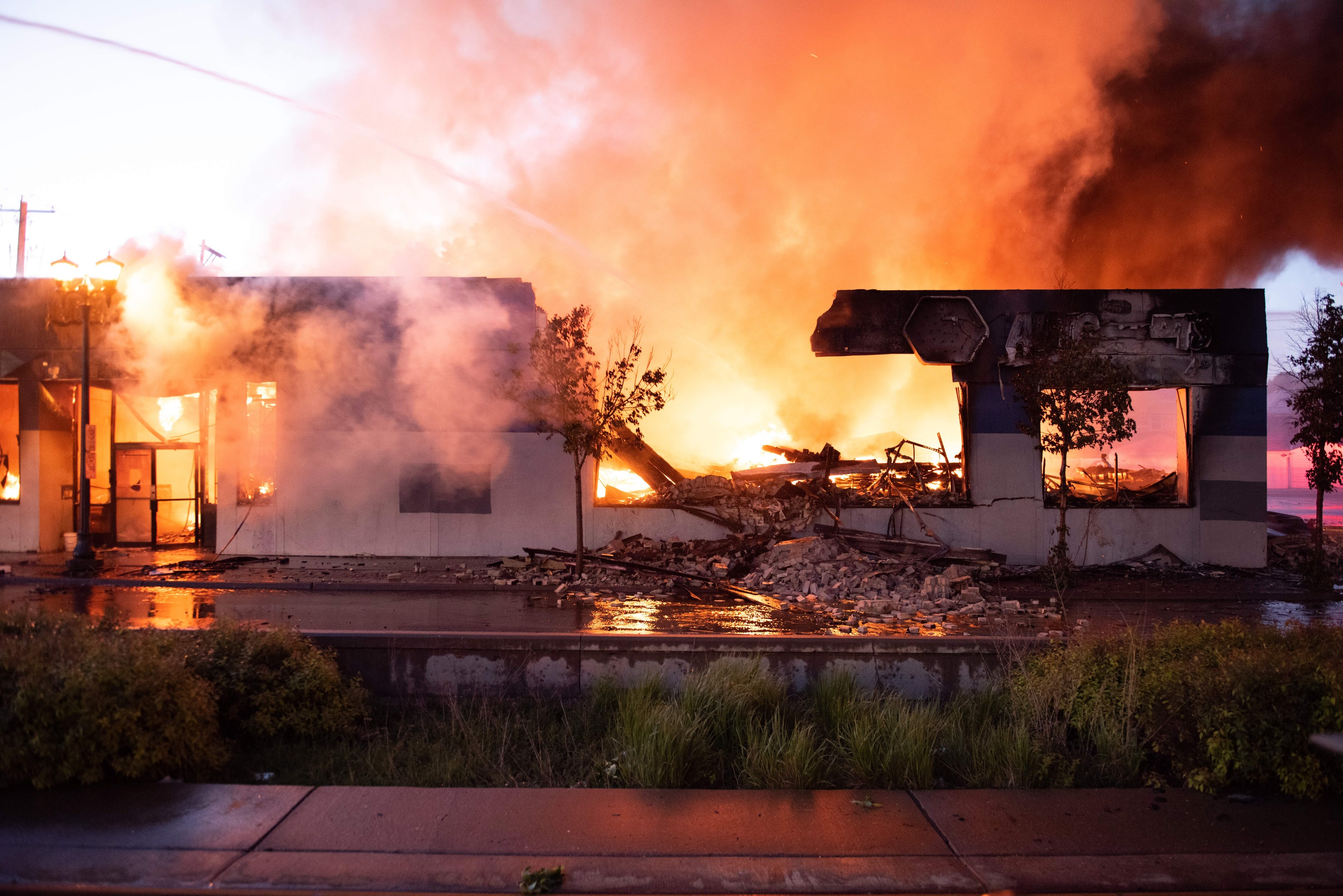 Haus in Flammen auf einer Straßenecke, mit dicken Rauchwolken, umgeben von Bäumen und Gras, und eine Straße unten sichtbar.