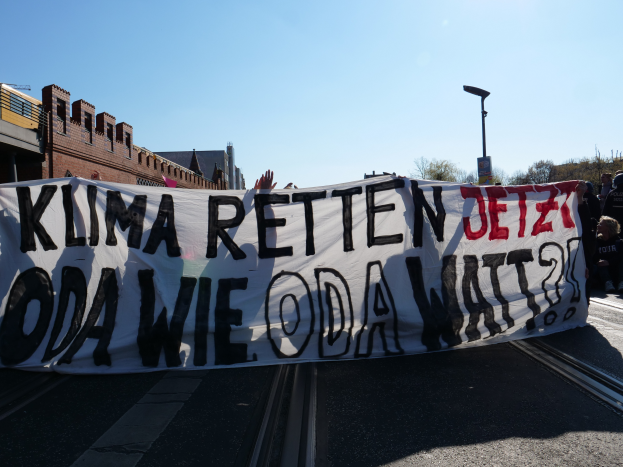 Eine Gruppe von Menschen steht an der Straße und hält ein Banner mit der Aufschrift "Klima Retten Jetz Oda Wie Oda Watt?" mit Gebäuden, Bäumen und einem Laternenpfahl im Hintergrund.
