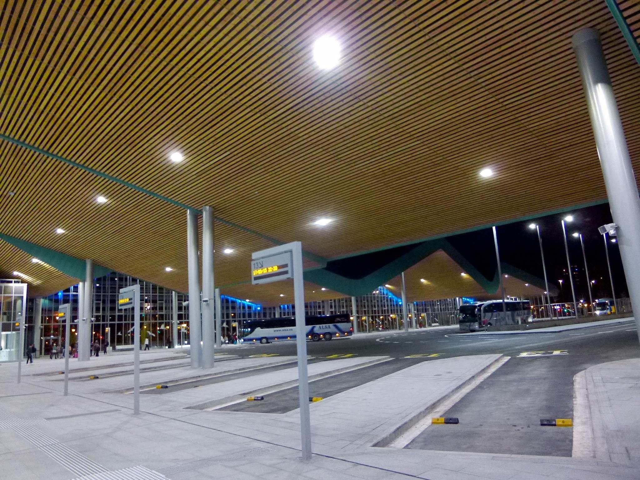 Eine Bushaltestelle nachts mit hellen Deckenleuchten, Fahrzeugen auf der Straße, Beschilderung, einigen Menschen und beleuchteten Gebäuden im Hintergrund.