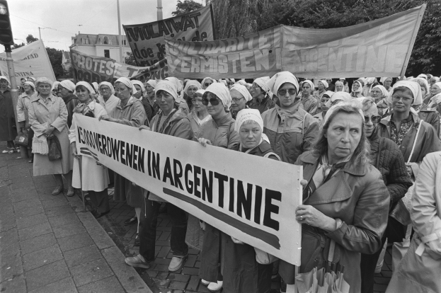Eine Gruppe von Frauen, die auf der Straße stehen und ein Banner halten, auf dem "Argentinischer Frauenmarsch in Argentinien" steht, mit Gebäuden, Bäumen und einem bewölkten Himmel im Hintergrund.
