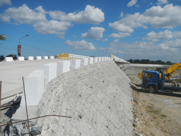 Baustelle mit einem Haufen Betonblöcke, einem Fahrzeug, einem Zaun, Steinen, Bäumen, einem Straßenpfahl, einem Schild und einem bewölkten Himmel.