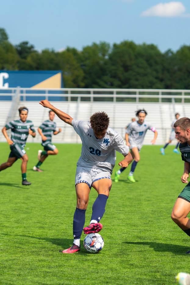 Eine Gruppe junger Männer, die Fußball auf einem Feld mit Bäumen und einem klaren blauen Himmel spielt, wobei ein Spieler Turnschuhe trägt.