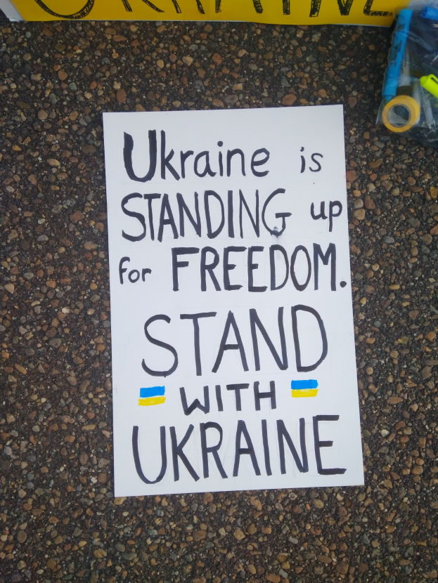 Ein weißes Papier mit dem Text "Ukraine steht für die Freiheit auf, stellt dich mit Ukraine" liegt vor einer gelben Flagge auf dem Boden, mit einer Plastikhülle mit verschiedenen Gegenständen rechts.