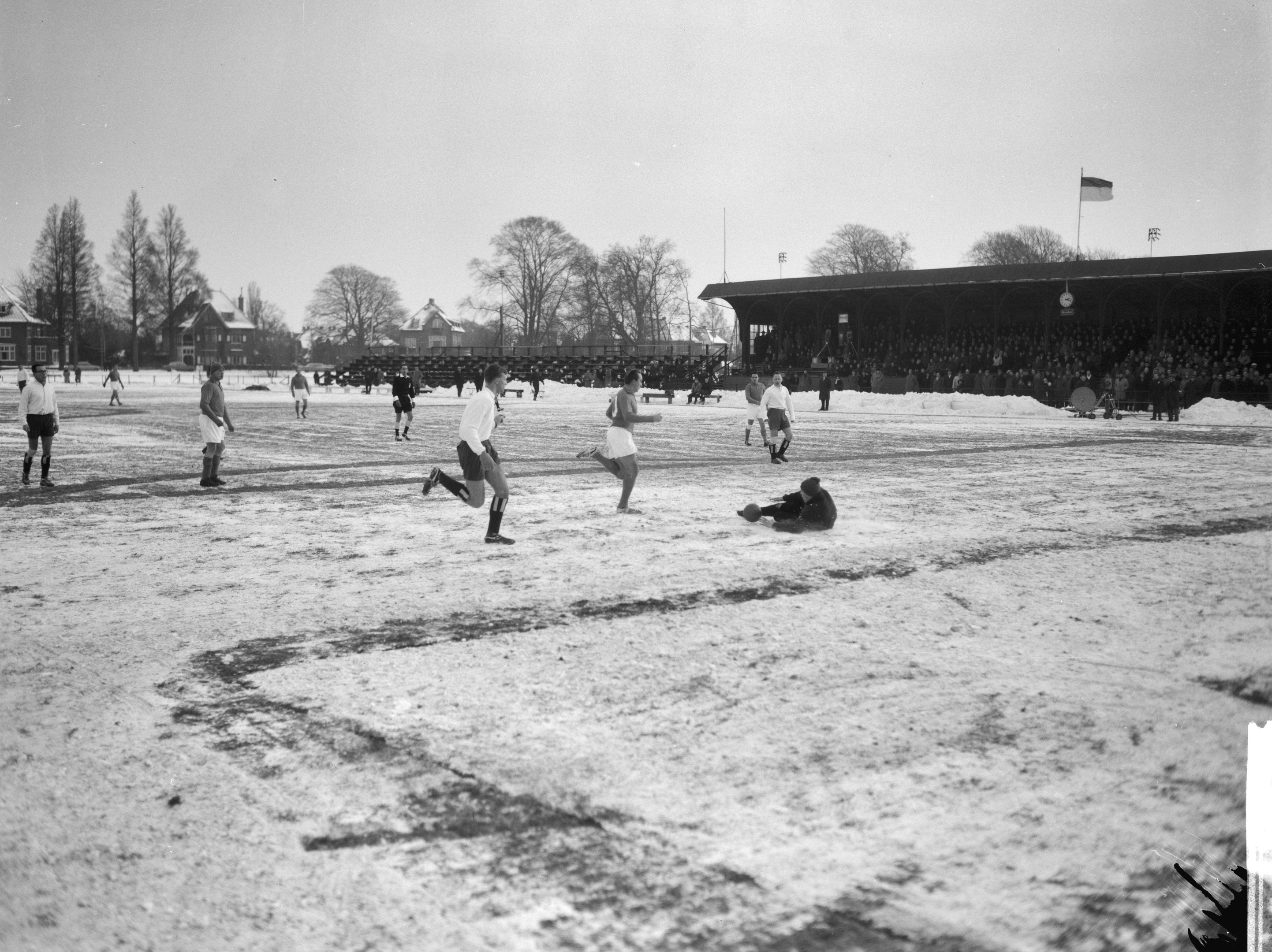 Schwarzes und weißes Bild von Menschen, die auf einem schneebedeckten Feld Fussball spielen, mit Häusern, Bäumen, Pfählen, Flaggen und einer Hütte im Hintergrund.