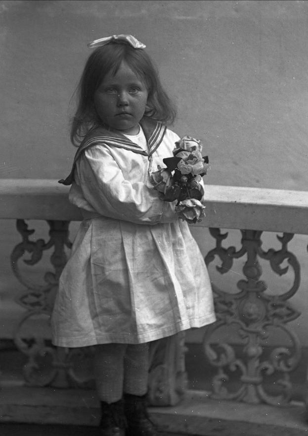Ein Schwarz-Weiß-Foto eines jungen Mädchens, das auf einem Balkon steht und einen Blumenstrauß hält, mit einem Geländer und einer Wand dahinter.