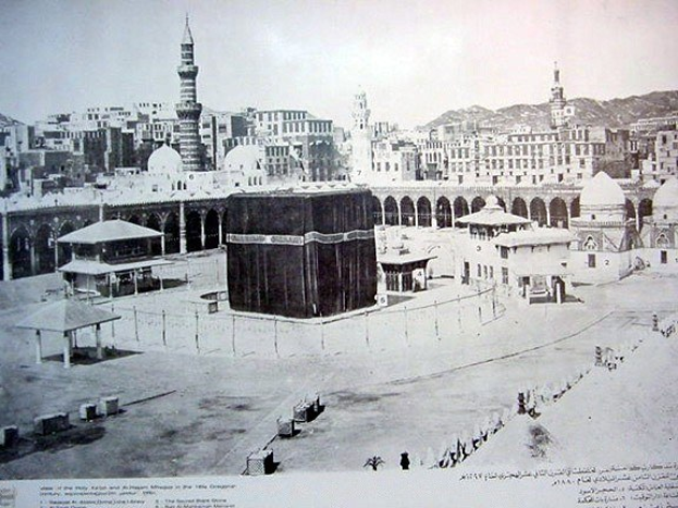Schwarzes und weißes Foto der Kaaba in Mekka, Saudi-Arabien, ein quadratischer Bau, der von einem Zaun umgeben ist, mit Menschen davor, vor einem Hintergrund aus Gebäuden, Bergen und einem klaren Himmel, mit Text unten.