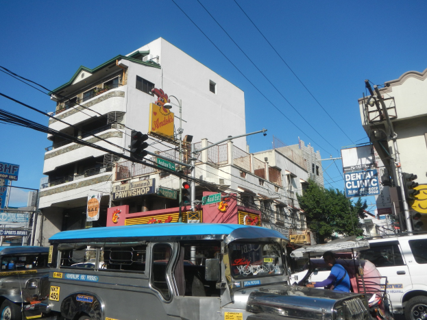 Eine belebte Straße in Manila, Philippinen mit Jeepneys, anderen Fahrzeugen, Füpfern, Gebäuden, Verkehrsampeln, Versorgungsmasten, Oberleitungen, Bäumen und einem klaren blauen Himmel.