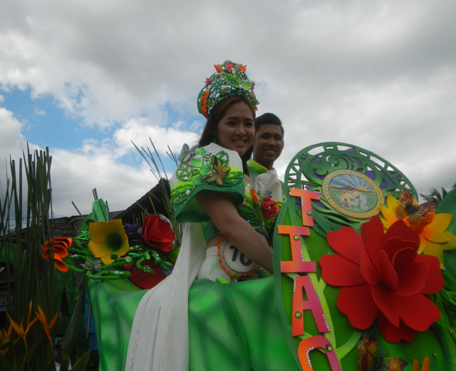 Ein Mann und eine Frau auf einem grünen und weißen Paradewagen, wobei die Frau ein grünes Kleid trägt und der Mann hinter ihr steht; der Wagen ist mit Blumen und Text geschmückt, vor einem Hintergrund aus Bäumen und einem klaren blauen Himmel.