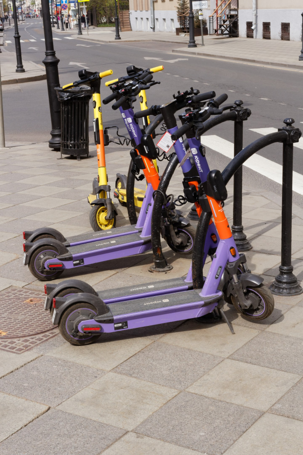 Eine Reihe von E-Scootern, die auf einer Straße geparkt sind, mit einem Müllcontainer im Vordergrund und Gebäuden, Bäumen und Fahrzeugen im Hintergrund.