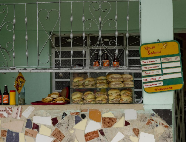 Ein Tablett mit Burgern, Flaschen und Tafeln mit Text, mit Grills im Hintergrund, einer Wand, einem Fenster und einer Säule auf der rechten Seite.