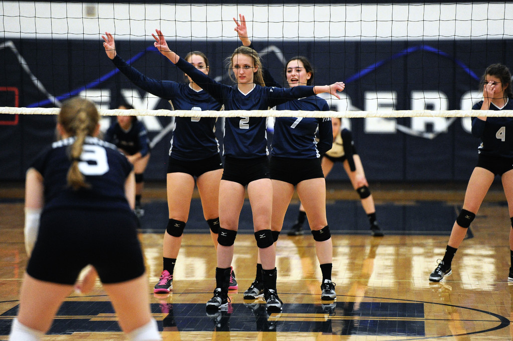 Drei Mädchen bei einem Volleyballspiel auf dem Feld stehend, mit erhobenen Händen, einem Netz davor und einem Zaun im Hintergrund.