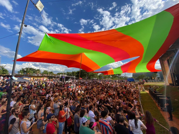 Eine große Menschenmenge steht vor einer prominenten Regenbogenflagge, mit einem Gebäude auf der rechten Seite, Lautsprechern auf der Bühne und Bäumen vor einem bewölkten Himmel, die sich zu einem Festivaljubiläum versammelt haben.