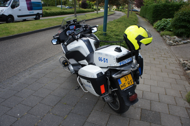Ein Polizei-Motorrad am Straßenrand geparkt mit einem Helm darauf, umgeben von Bäumen, Polen, Gebäuden, Pflanzen, Gras, Steinen und anderen Fahrzeugen im Hintergrund.