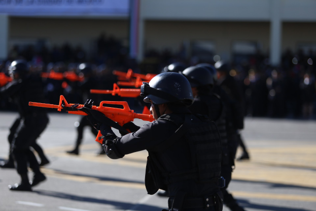 Polizisten in Einsatzausrüstung marschieren auf einer Straße, tragen Helme und halten Waffen, mit einem Gebäude und einer Fahne im Hintergrund.