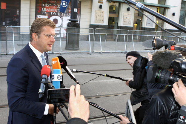 Ein Mann in Anzug steht vor Reportern mit Kameras und Mikrofonen, mit einem Metallzaun, einer Stange, einem Müllcontainer, Schildern, einem Gebäude und angehefteten Papieren im Hintergrund.