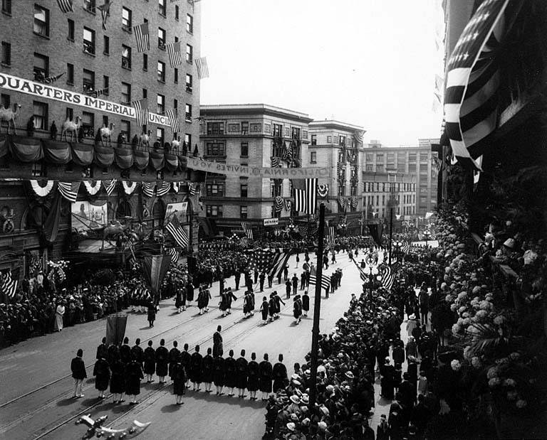 Eine große Menschenmenge marschiert auf einer Straße in New York City während eines Fourth-of-July-Umzugs, viele tragen Mützen und halten Fahnen, während Zuschauer an den Seiten und Gebäude im Hintergrund stehen.