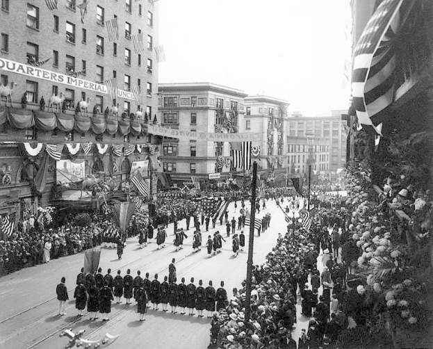 Eine große Menschenmenge marschiert auf einer Straße in New York City während eines Fourth-of-July-Umzugs, viele tragen Mützen und halten Fahnen, während Zuschauer an den Seiten und Gebäude im Hintergrund stehen.