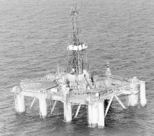 Schwarz-weiß-Foto einer Ölplattform in der Mitte des Ozeans, umgeben von Wasser und einer fernen Brücke mit Säulen und einem Turm.