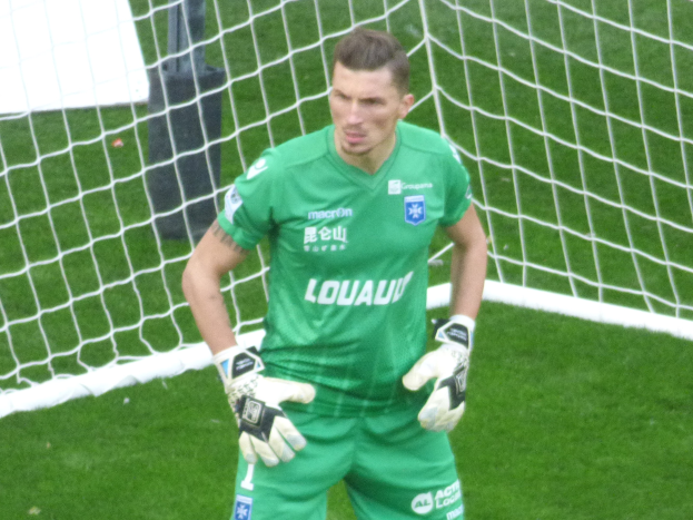 Ein Torwart in grüner Uniform steht vor einem Fußballtor und trägt Handschuhe auf einem grünen Rasen.