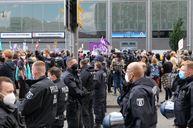 Große Gruppe von Menschen protestiert vor einem Gebäude, einige halten Schilder und tragen Helme, mit einem Schildständer im Vordergrund und einem Baum im Hintergrund.