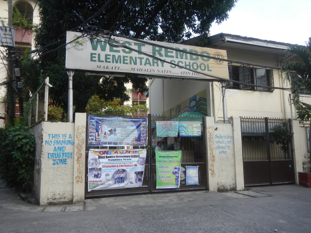 Außenansicht der West Rembo-Grundschule in Manila, Philippinen, die ein Gebäude mit Fenstern, ein Tor, eine Schautafel, Plakate, Pflanzen, Bäume, einen Straßenmast, Drähte und einen bewölkten Himmel zeigt.