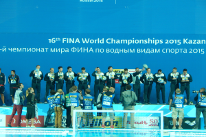 Eine Gruppe von Menschen steht oben auf einem Swimmingpool, einige halten Kameras, mit einem Netz im Vordergrund und einer Fahne im Hintergrund, die den Text 'FINA World Championships 2015 Kazan' trägt.