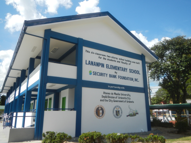 Ein zweistöckiges Schulgebäude mit weißen Wänden, Säulen, Geländern und Fenstern, umgeben von Grün, geparkten Fahrzeugen, Strommasten und einem bewölkten Himmel.