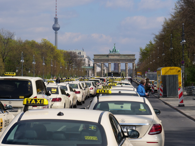 Eine belebte Straße in Berlin, Deutschland, mit vielen parkenden Taxis, Fußgängern auf dem Gehweg, Laternenpfählen, Bäumen und Gebäuden im Hintergrund, mit einem fernen Bogen mit Statuen und einem Turm unter einem bewölkten Himmel.