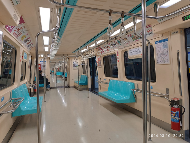 Innenseite eines U-Bahn-Wagens mit blauen Sitzen, Griffen, Stäben, Deckenleuchten, Fenstern, Plakaten und einem Feuerlöscher; mehrere Fahrgäste sind sitzend zu sehen, was eine aktive Stadtumgebung wiederspiegelt.