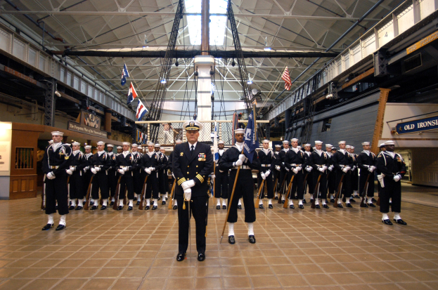 Eine Gruppe uniformierter Männer in einem großen Raum mit Waffen, Fahnen, Texttafeln an den Wänden und Deckenleuchten und Stangen.