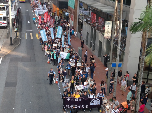 Eine große Gruppe von Menschen marschiert auf einer Straße in Hong Kong, wobei sie Protestplakate und Schilder halten und Bäume, Glasfassaden, Fahrzeuge und Schilder sichtbar sind.