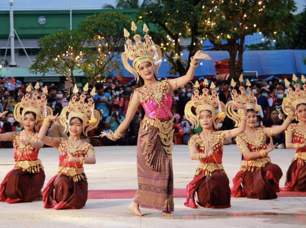Eine Gruppe von Frauen in traditioneller Thai-Kleidung, die vor einem sitzenden und stehenden Publikum tanzen, mit Bäumen, Lichtern, Pfosten, einem Gebäude und einem klaren blauen Himmel im Hintergrund.
