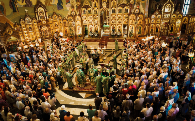 Eine große Gruppe von Menschen steht vor einer russisch-orthodoxen Kirche und hält Musikinstrumente, Kerzen auf Ständern, einen Teppichboden, Fotorahmen an den Wänden und ein Fenster auf der rechten Seite.