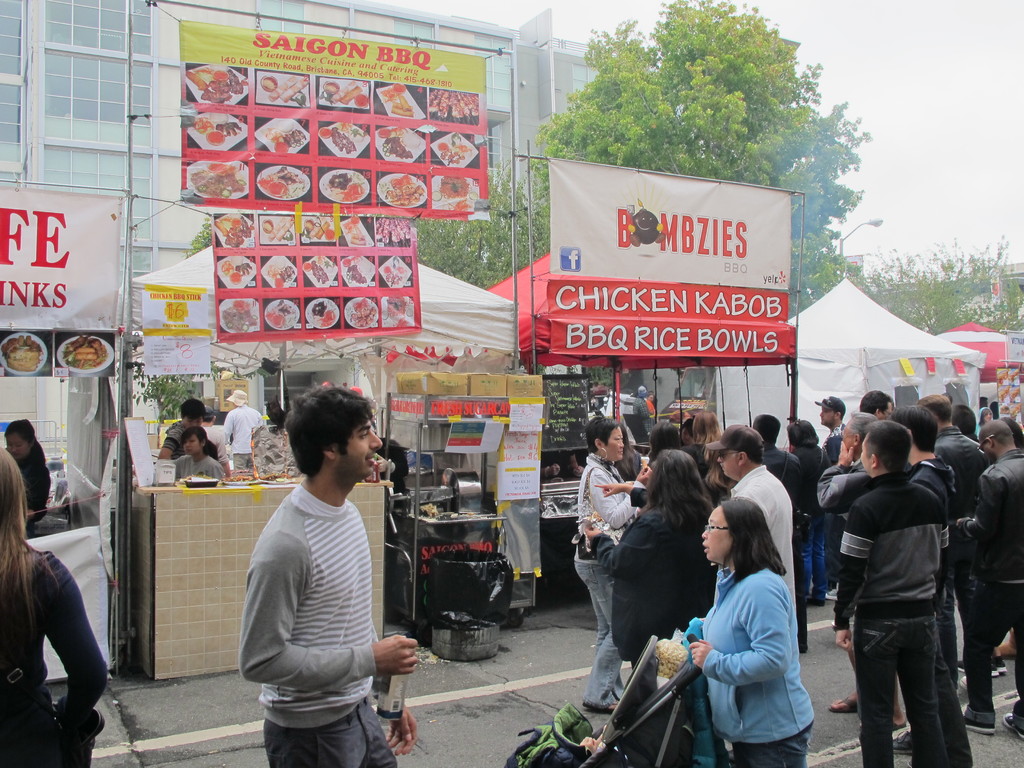 Menschen auf einer Straße außerhalb der Stadt, mit einem Mann in einem grauen und weißen T-Shirt, der eine Wasserflasche und eine Frau in einer blauen Jacke hält, die einen Kinderwagen schiebt. Hinter ihnen sehen wir Stände mit weißen und roten Zelten, farbigen Bannern und Gebäuden, Bäumen und einer Straßenlaterne im Hintergrund.