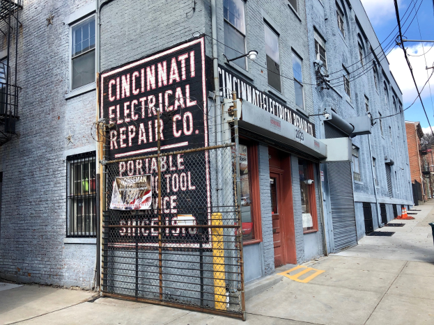 Außenansicht des Cincinnati Electrical Repair Co. Gebäudes mit Fenstern, Türen, einem Schild, einem Metallzaun, einem Müllcontainer, Strommasten, Drähten, Bäumen und einem bewölkten Himmel.