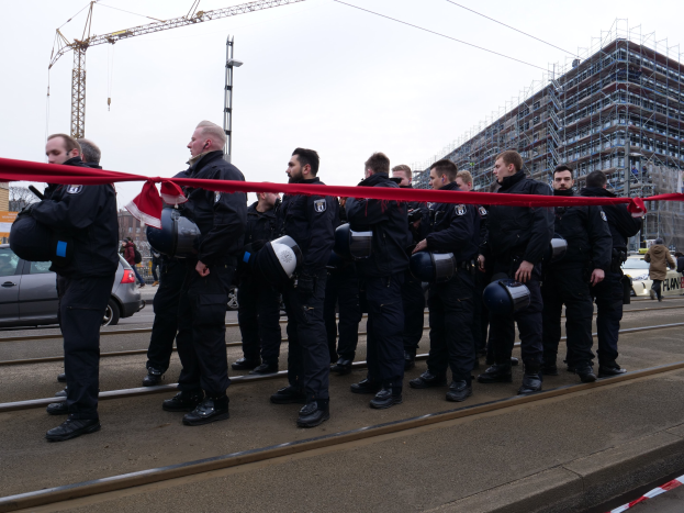 Eine Gruppe von Polizisten in schwarzen Uniformen und Helmen steht in einer Reihe auf einem Bahngleis, mit einem roten Band davor, während im Hintergrund Fahrzeuge, Fußgänger, Gebäude, Strommasten und ein klarer blauer Himmel zu sehen sind.