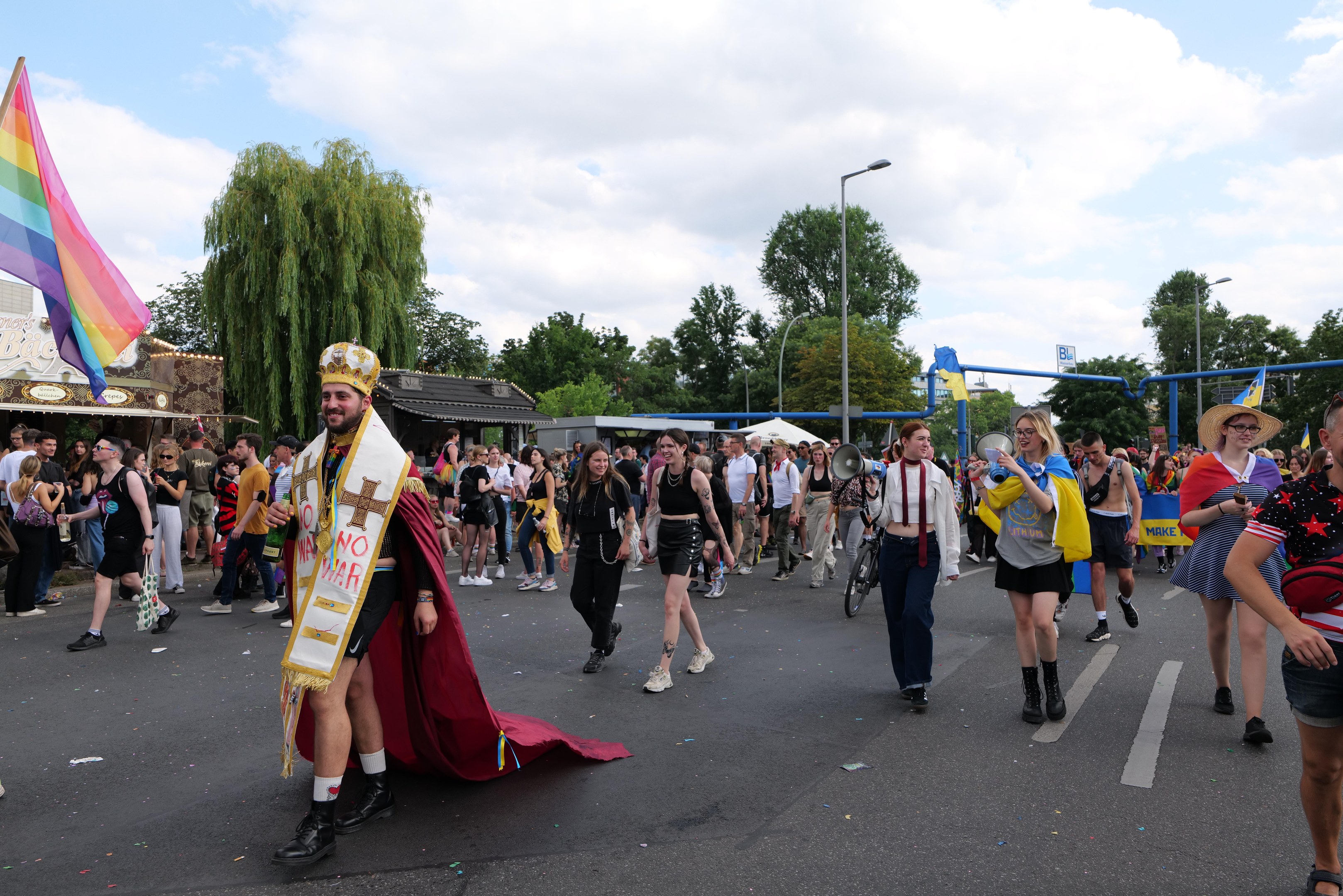 Eine Gruppe von Menschen marschiert bei der Pride Parade 2018 mit einer Regenbogenflagge und Musikinstrumenten, umgeben von Laternenpfählen, Bäumen, Hütten und einem bewölkten Himmel.