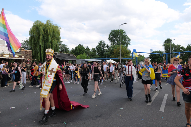 Eine Gruppe von Menschen marschiert bei der Pride Parade 2018 mit einer Regenbogenflagge und Musikinstrumenten, umgeben von Laternenpfählen, Bäumen, Hütten und einem bewölkten Himmel.