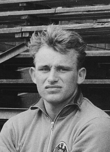 Schwarzes und weißes Foto eines Mannes in einer Jacke, der vor einem Stapel hölzerner Planken steht, der als legendärer deutscher Fussballer gilt.