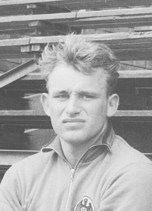 Schwarzes und weißes Foto eines Mannes in einer Jacke, der vor einem Stapel hölzerner Planken steht, der als legendärer deutscher Fussballer gilt.