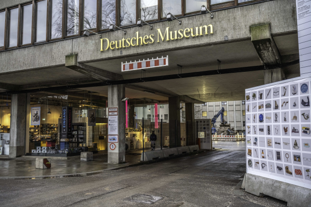 Außenansicht des Deutschen Museums in Berlin, mit einem großen Glasgebäude mit Säulen, einem Namensschild, einer Anzeigetafel mit Bildern und Text sowie umliegenden Bäumen und anderen Gebäuden vor einem Himmel-Hintergrund.