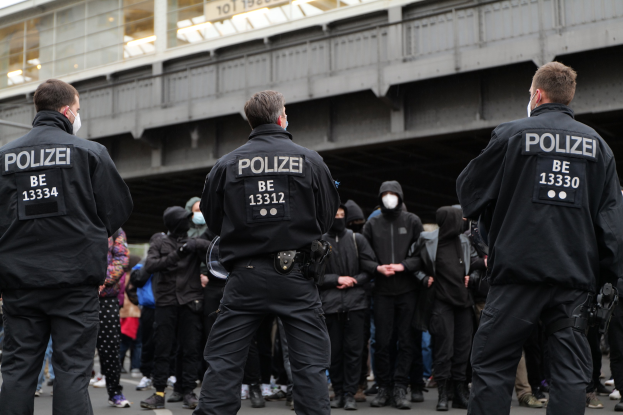 Polizeibeamte in schwarzen Uniformen und Masken stehen vor einer Menge, mit einer Brücke und einem Gebäude im Hintergrund.