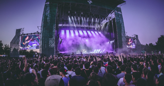 Eine große Menschenmenge bei einem Musikfestival schaut auf einer beleuchteten Bühne mit Instrumenten und Bildschirmen, die vor einem Gebäude, Bäumen und einem blauen Himmel aufgebaut ist.