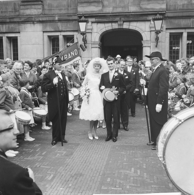Ein Brautpaar geht eine Straße entlang vor einer Menge, einige halten Musikinstrumente, mit einem Gebäude und einer Fahne im Hintergrund, alles in Schwarz-Weiß dargestellt.