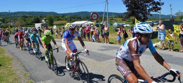 Eine Gruppe von Radfahrern fährt eine Straße entlang, während Zuschauer am Straßenrand stehen, einige halten Fahnen und tragen Helme oder Schutzbrillen, mit Bäumen, Fahrzeugen, Zelten und Gebäuden im Hintergrund unter einem klaren blauen Himmel.