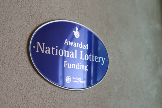 Blaues rechteckiges Schild an einem Gebäude mit weißem Hintergrund und schwarzer fetter Schrift, die "National Lottery Funding" lautet.
