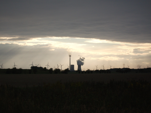 Kraftwerk mit Windrädern am wolkenverhangenen Himmel, umgeben von Grünzeug am Boden.