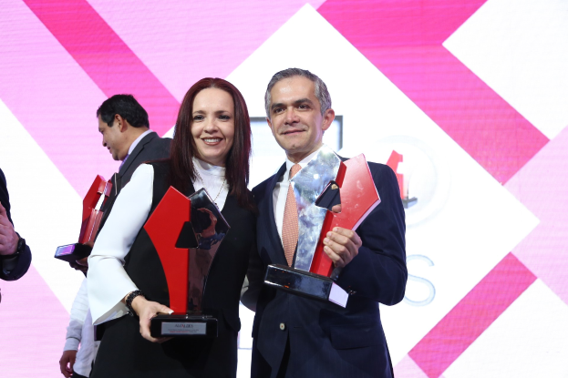 Zwei Personen auf einer Bühne, die lächeln und Trophäen halten, mit einem Bildschirm im Hintergrund.