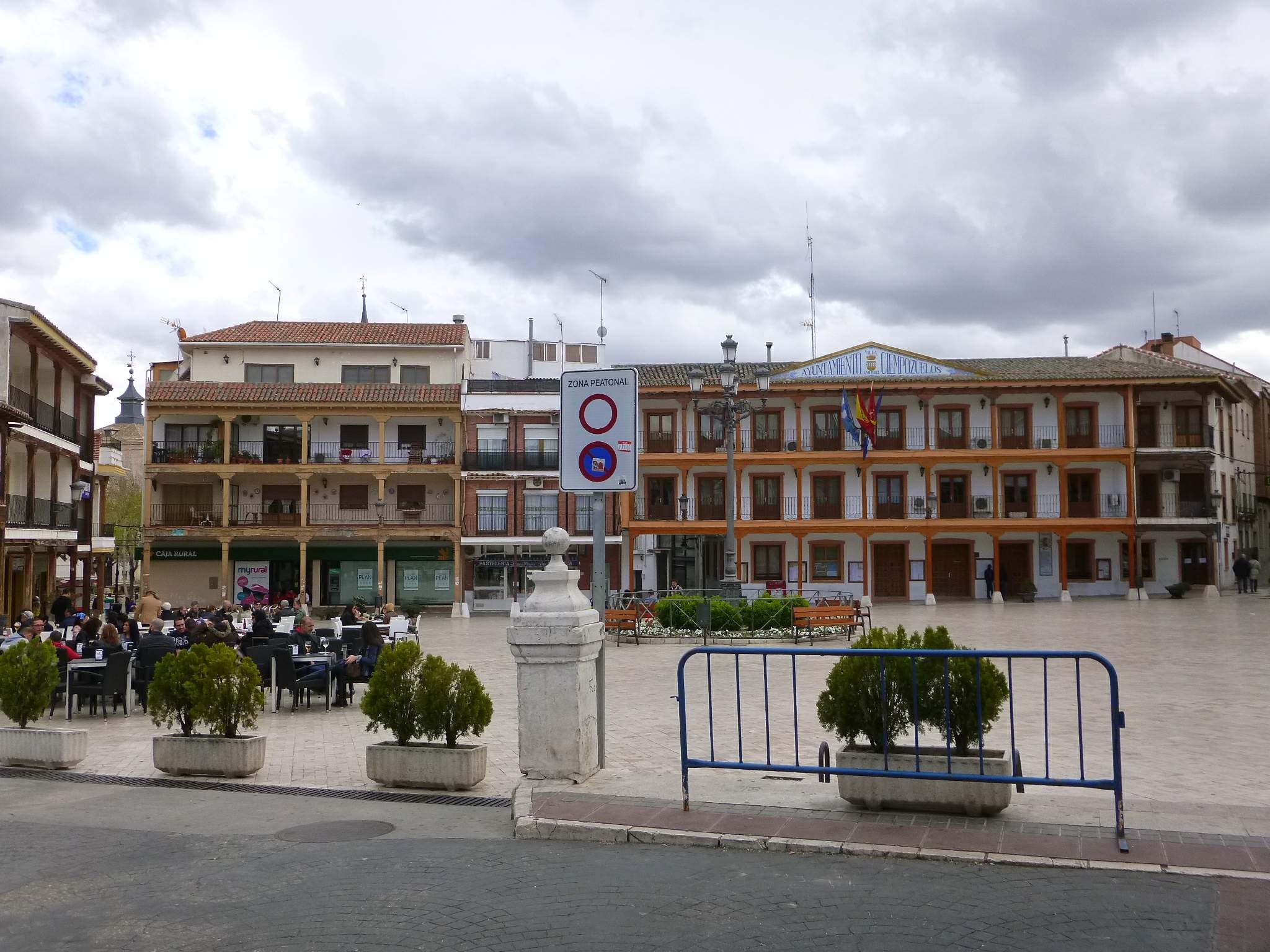 Ein belebter Plaza Mayor mit Menschen auf Stühlen sitzend und stehend, umgeben von Topfpflanzen, Metallabsperrungen, einem Schild an einem Pfahl, Straßenmasten mit Flaggen, Gebäuden mit Fenstern und einem bewölkten Himmel.