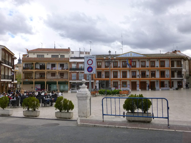 Ein belebter Plaza Mayor mit Menschen auf Stühlen sitzend und stehend, umgeben von Topfpflanzen, Metallabsperrungen, einem Schild an einem Pfahl, Straßenmasten mit Flaggen, Gebäuden mit Fenstern und einem bewölkten Himmel.